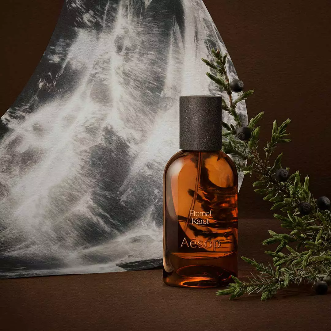 Amber Aesop fragrance bottle beside rosemary sprigs and a monochrome water image on a dark brown surface.